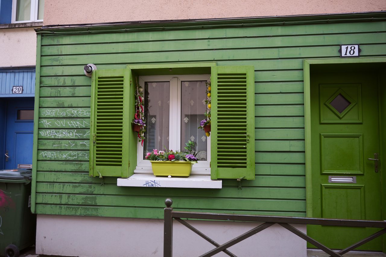 Façade de maison verte en été avec volets fermés, gouttières propres et jardin arrosé goutte-à-goutte.