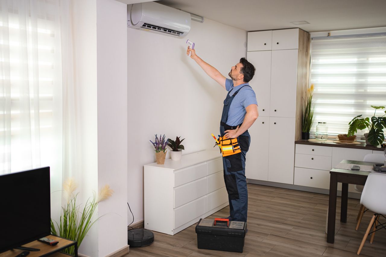 Technicien HVAC contrôlant la climatisation murale dans un salon lumineux avec thermomètre infrarouge et outils de maintenance.